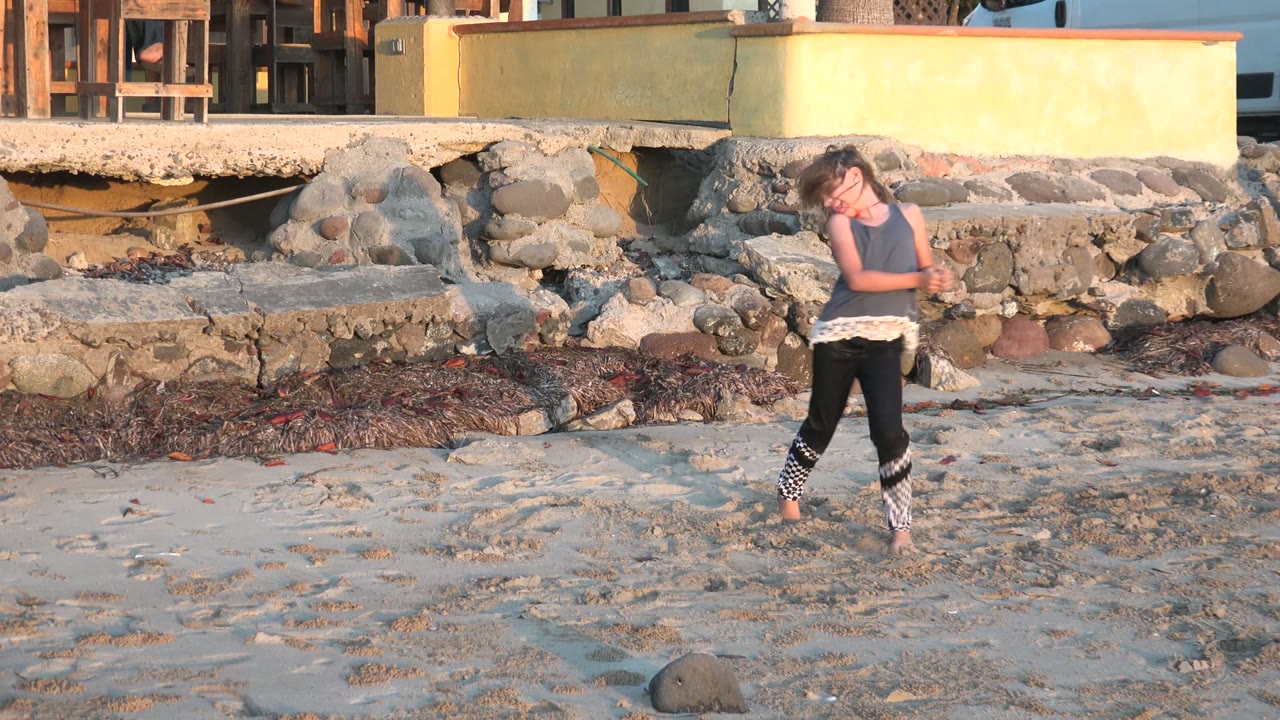 A girl dances barefoot on pale sand in San Carlos BCS Mexico, with her body angled toward camera and rocks, a wall, and beachside structures behind her.