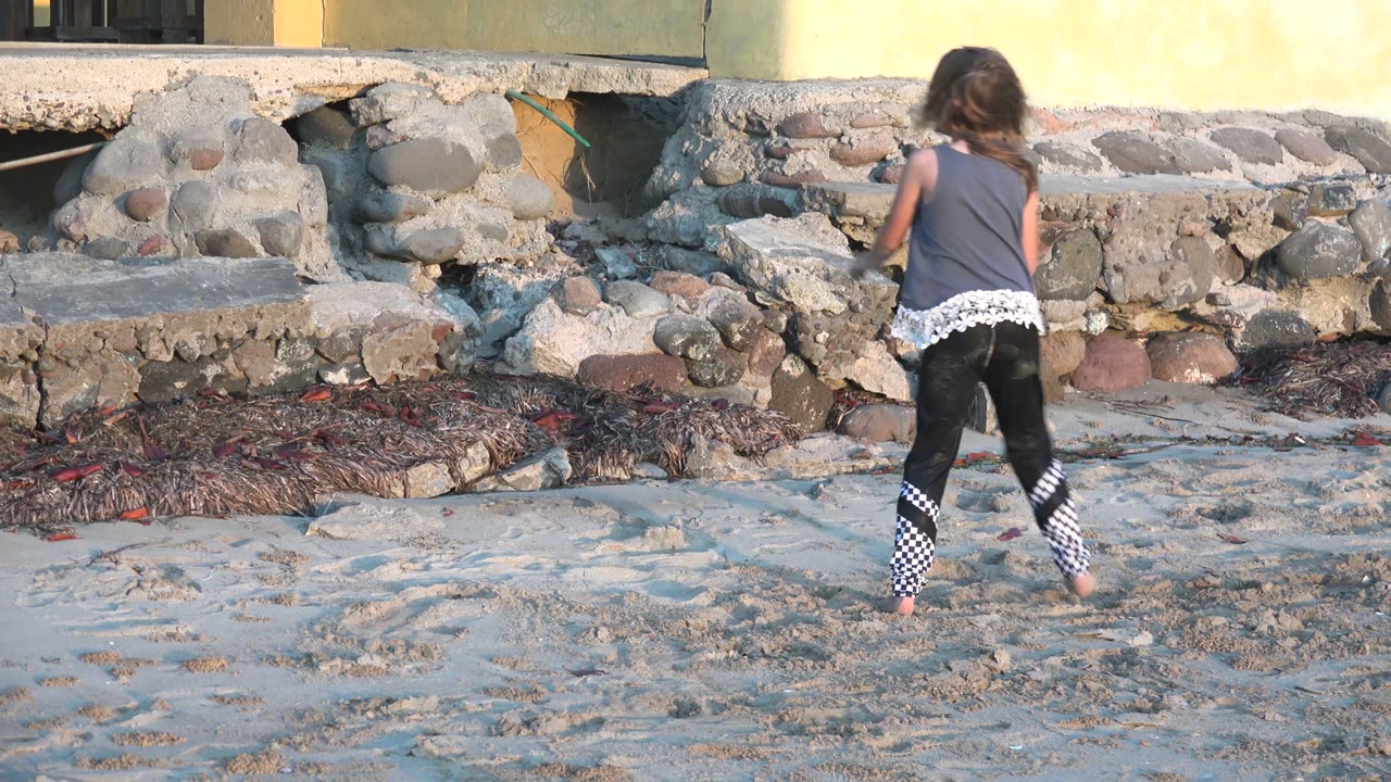 A girl seen from behind walks barefoot across sand near a rough stone wall in San Carlos BCS Mexico, with a yellow building wall behind her.