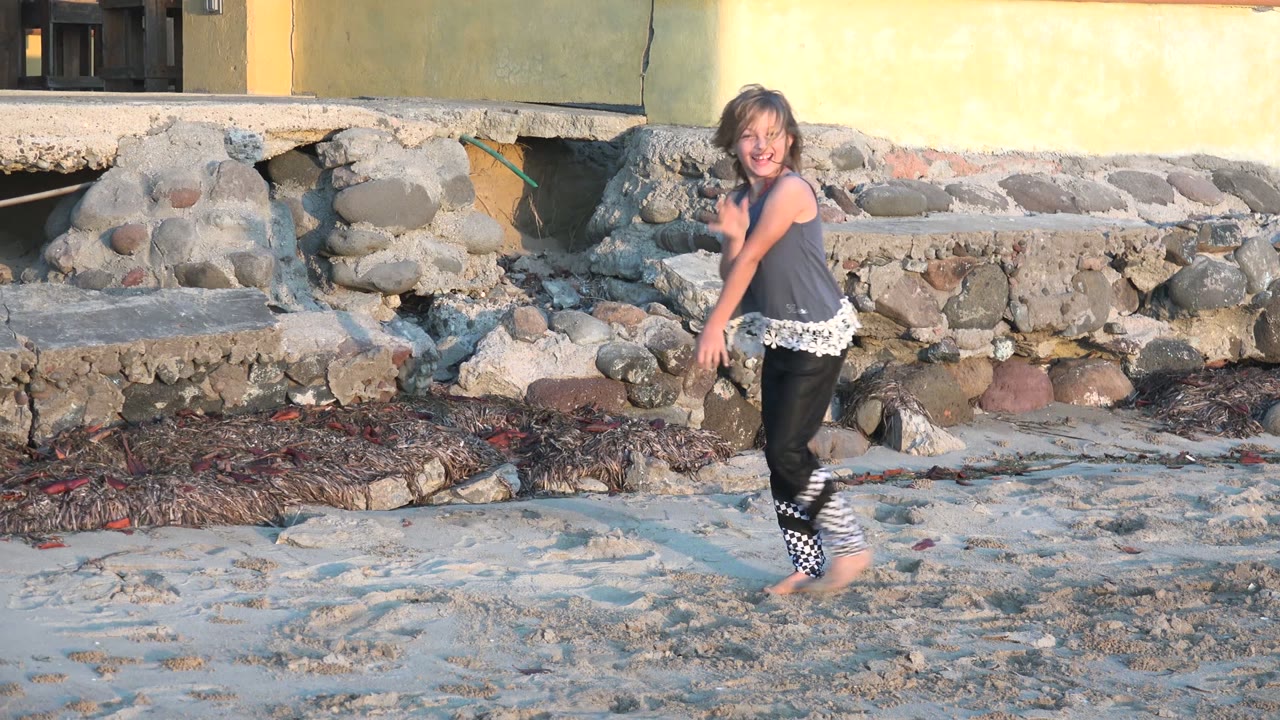 The girl faces partly toward camera while playing on the sand in San Carlos BCS Mexico, with her face visible and the stone wall behind her.
