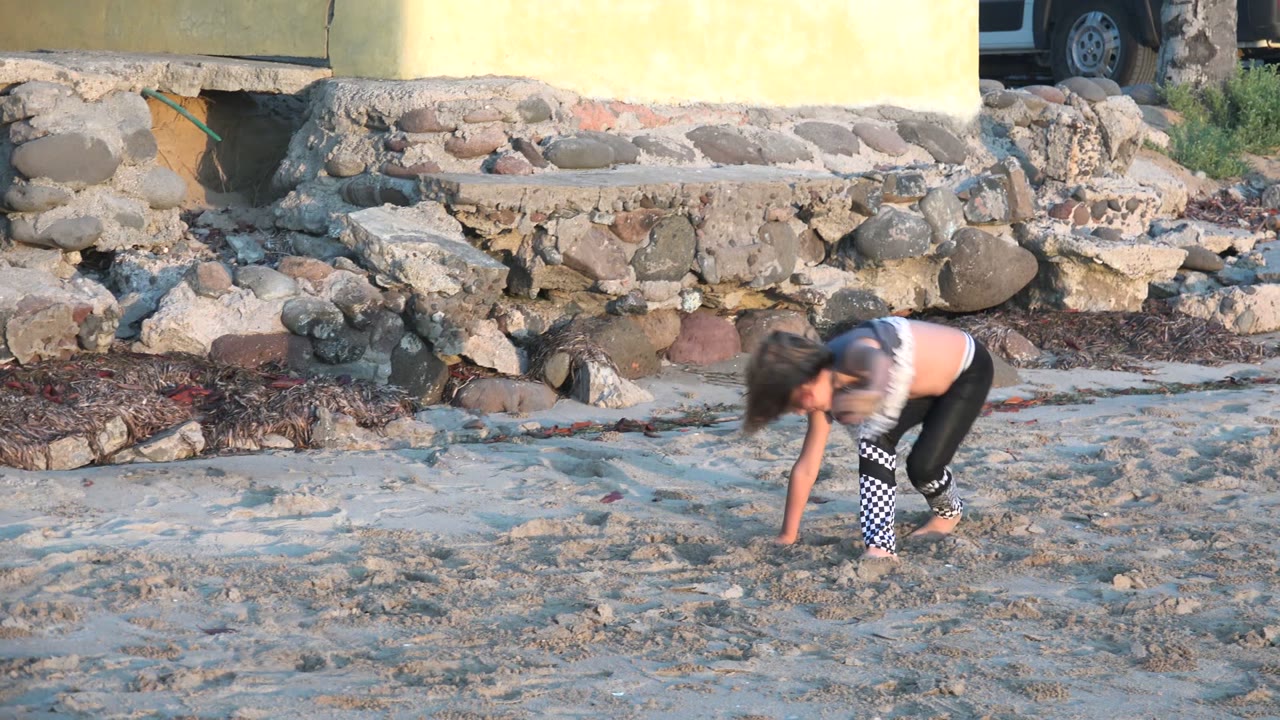 At 0:00 in San Carlos BCS Mexico, a child bends close to the sand near a rough stone wall, with rocks and a yellow building edge behind them.
