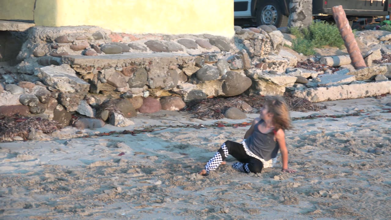 Child sitting on sand with one hand down near a rocky wall.