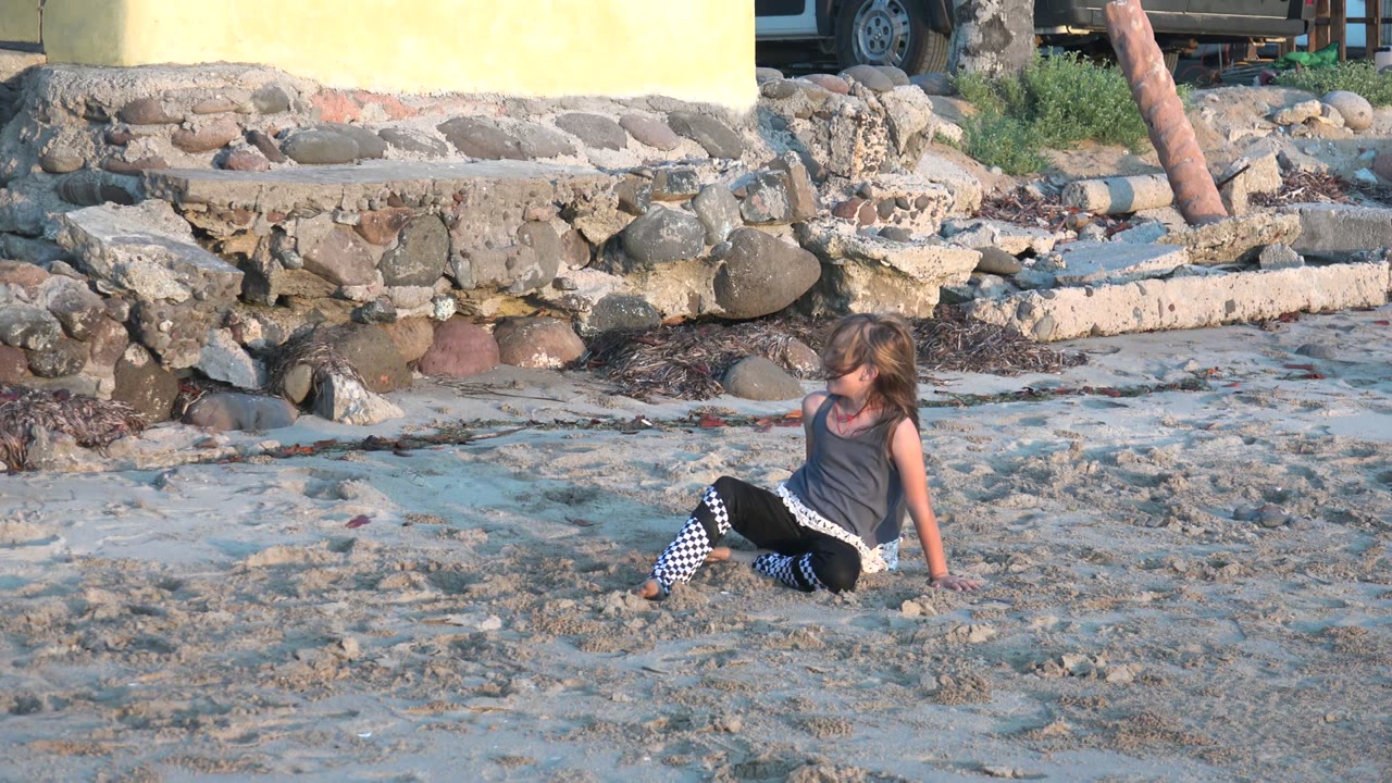 At 0:02 in San Carlos BCS Mexico, the child remains seated on the beach, turned slightly toward the sand, with rough stones, rubble, and a yellow wall in the background.