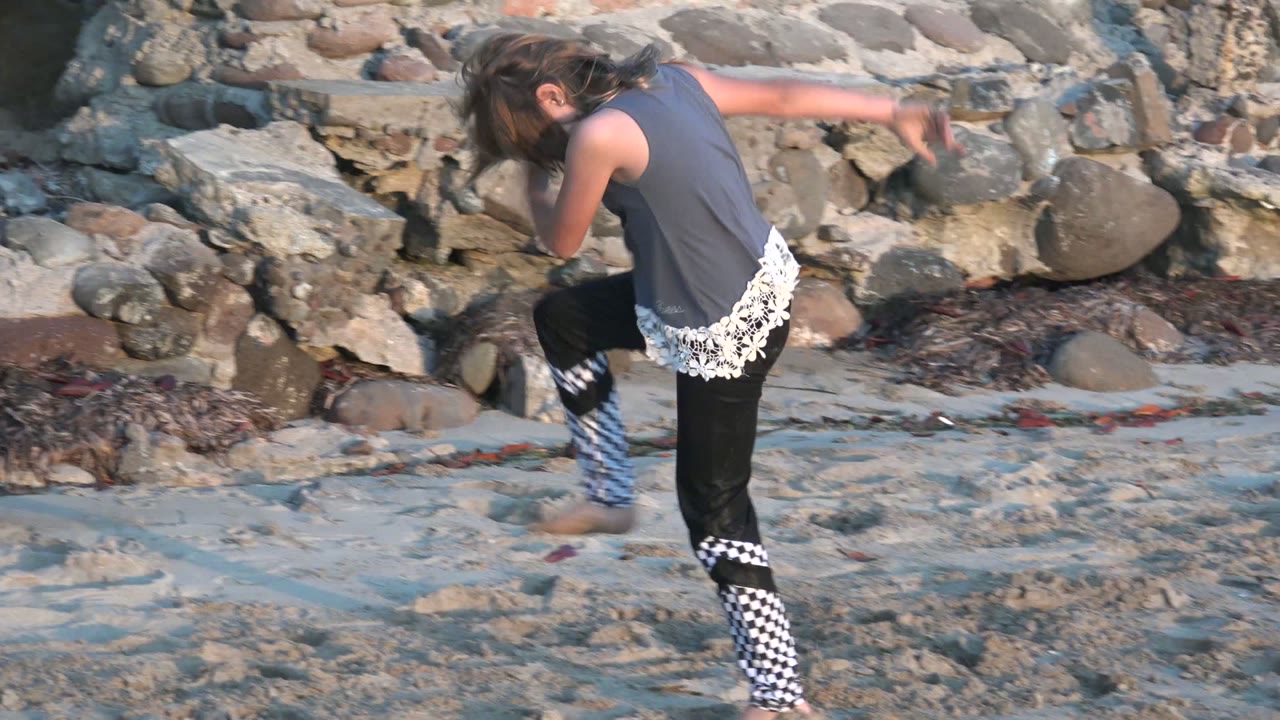 In San Carlos BCS Mexico, a child in a gray top and black pants lifts one leg on a sandy beach beside a rough stone seawall.