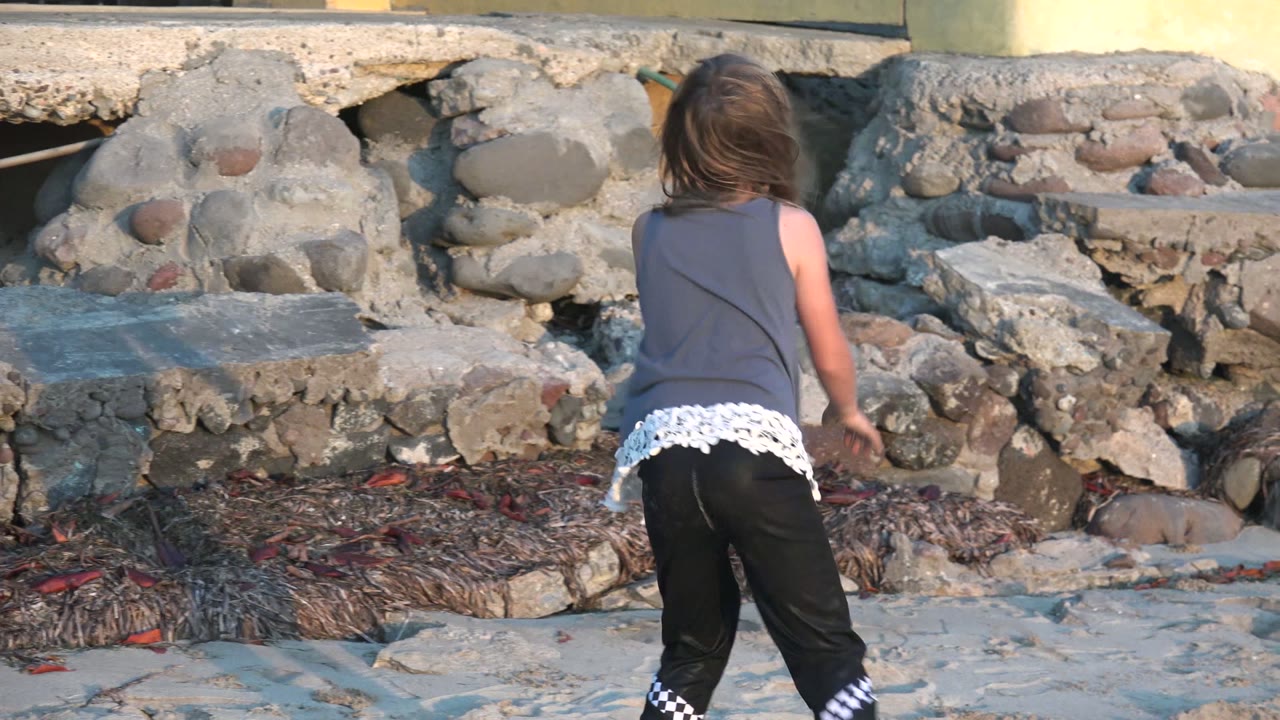 In San Carlos BCS Mexico, the child stands turned away from camera near the sandy edge, with a low rocky seawall and scattered beach debris behind her.