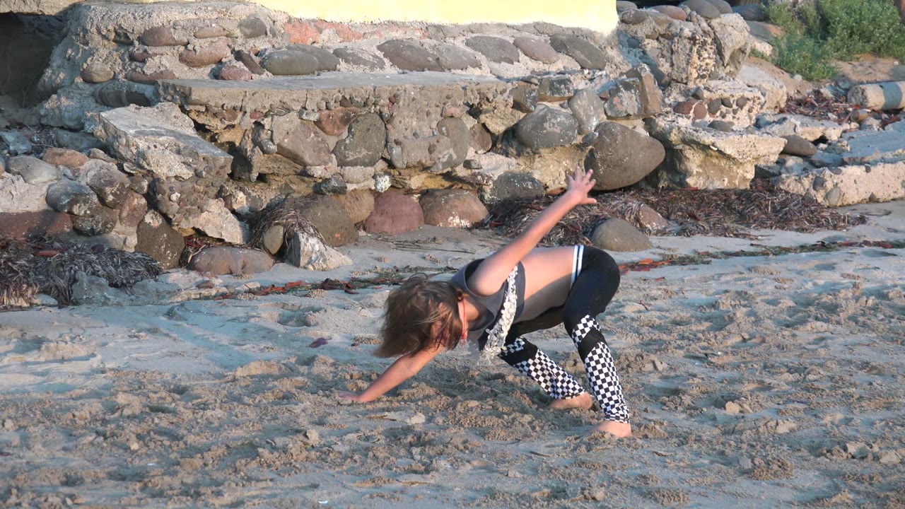 A girl crouches low with one arm raised while dancing barefoot on rippled sand in San Carlos BCS Mexico, with rocks and a yellow wall behind her.