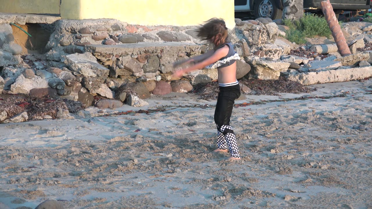 A girl rises out of a crouched dance position on the sandy shore in San Carlos BCS Mexico, with stones, a wall, and part of a parked vehicle visible behind her.
