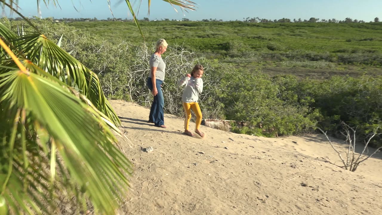 The woman and child continue across the sandy rise in San Carlos BCS Mexico, with the child slightly ahead and green vegetation filling the background.