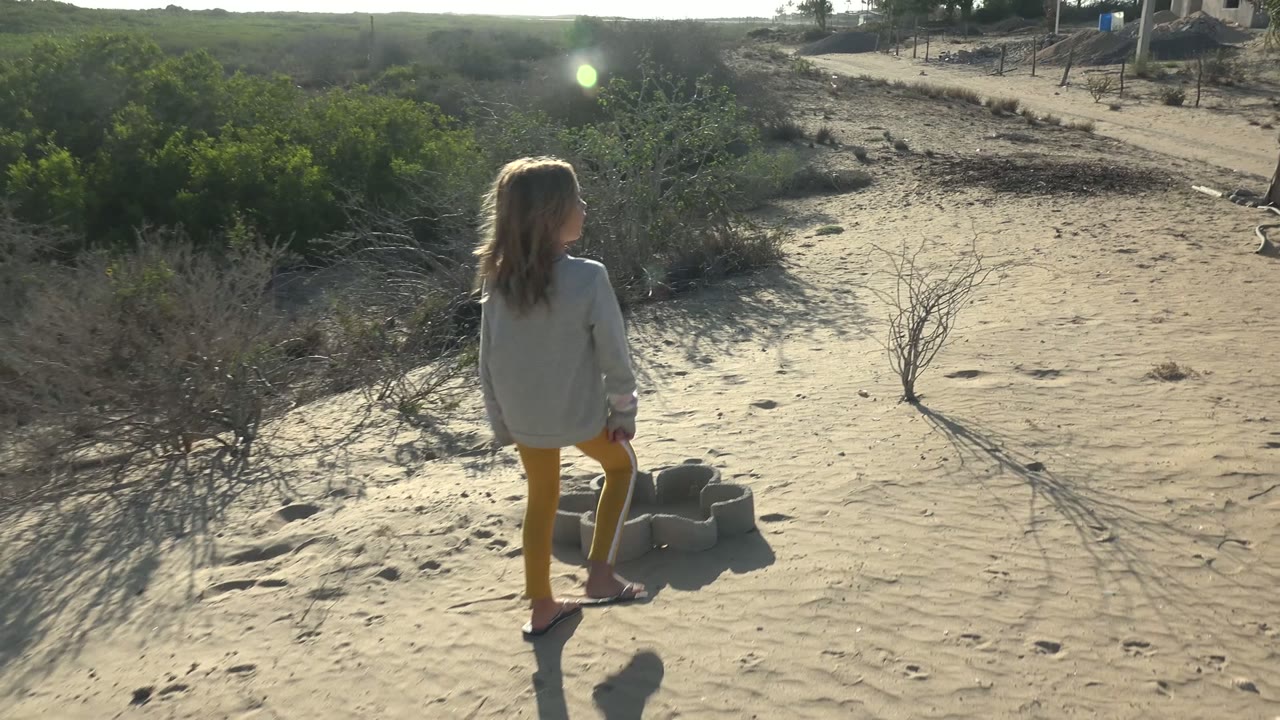A young girl stands on pale sandy terrain in San Carlos BCS Mexico, with small cinder blocks near her feet, low green shrubs, dry brush, and long shadows.