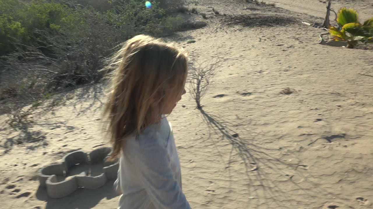 A close side view shows the young girl moving through sandy San Carlos BCS Mexico terrain, with dry brush, footprints, and desert plants around her.