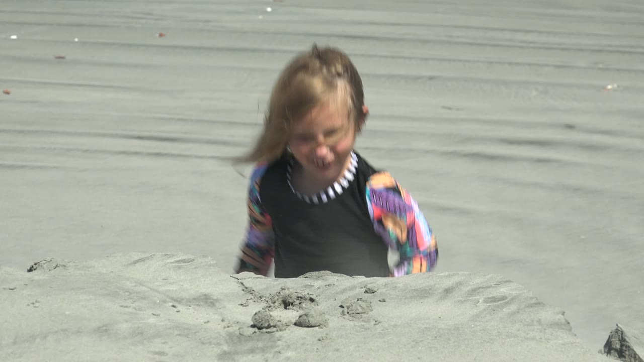 At La Bocana BCS Mexico, a young girl in a colorful long-sleeve beach shirt appears partly blurred behind a pale sand mound as she begins shaping the sand.