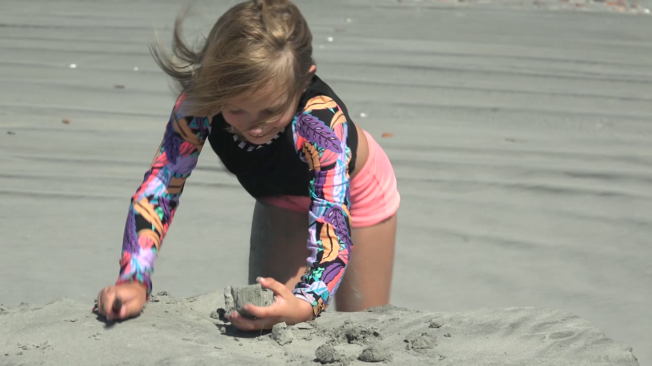 At La Bocana BCS Mexico, the young girl leans over the foreground sand mound with windblown hair, one hand holding sand and the other pressing into the beach.