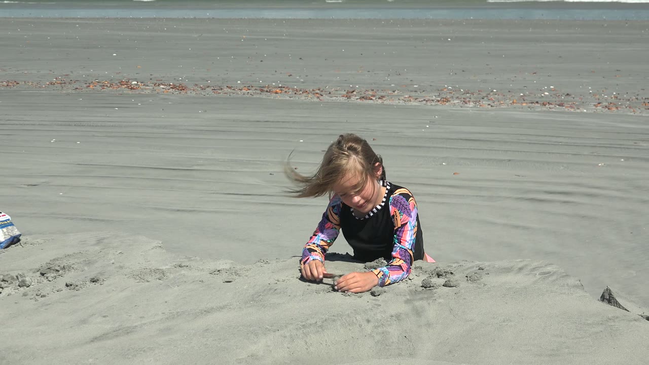 At La Bocana BCS Mexico, a young girl leans over pale beach sand near the foreground while white surf lines cross the distant ocean.