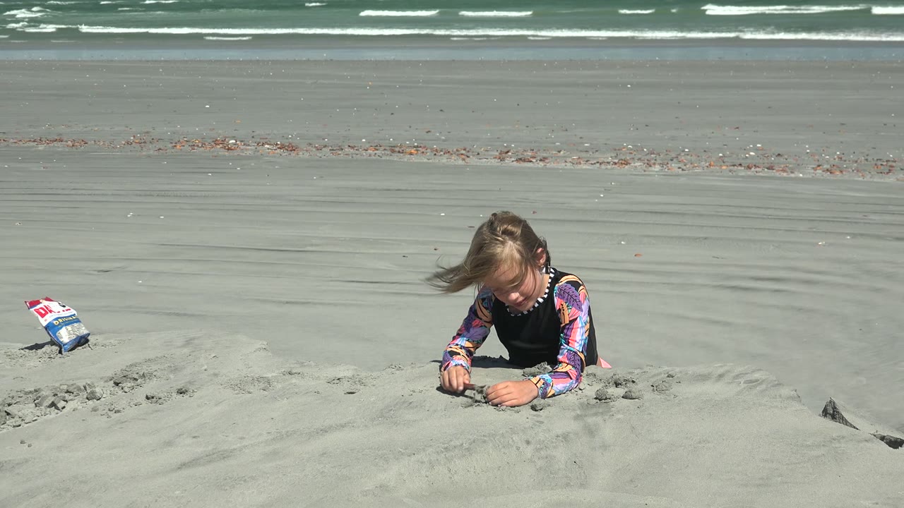 Girl in colorful sleeves works with sand beside a small bag on the beach.
