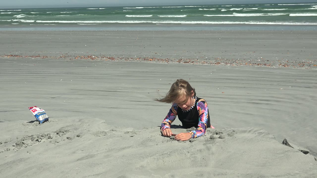 At La Bocana BCS Mexico, the child stays low behind the sand mound as wind pushes her hair and the ocean horizon remains steady behind her.