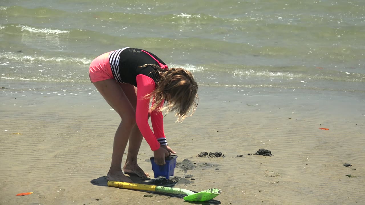 A young girl in a pink and black swimsuit bends over a blue sand bucket on the San Carlos BCS Mexico shoreline, with a green shovel and small waves behind her.