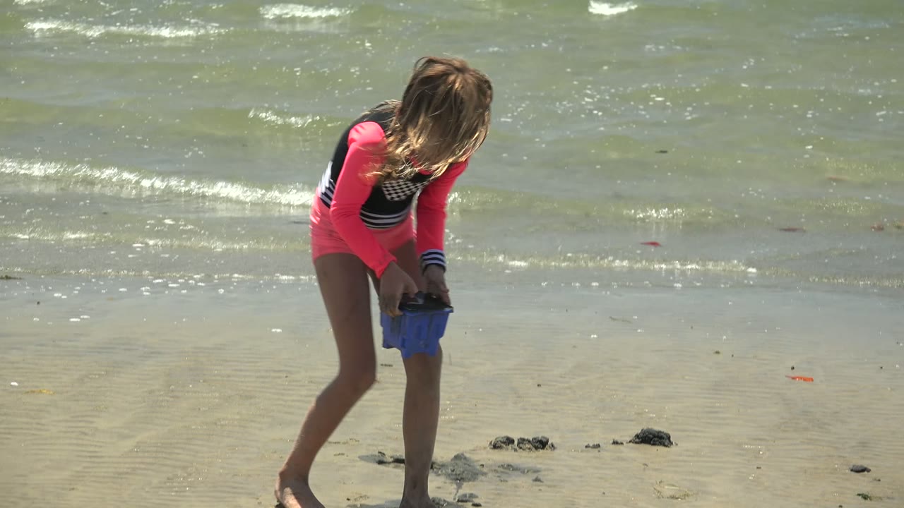 The girl stands on wet sand holding a blue bucket on the San Carlos BCS Mexico beach, with small rocks at her feet and wind-ruffled water in the background.