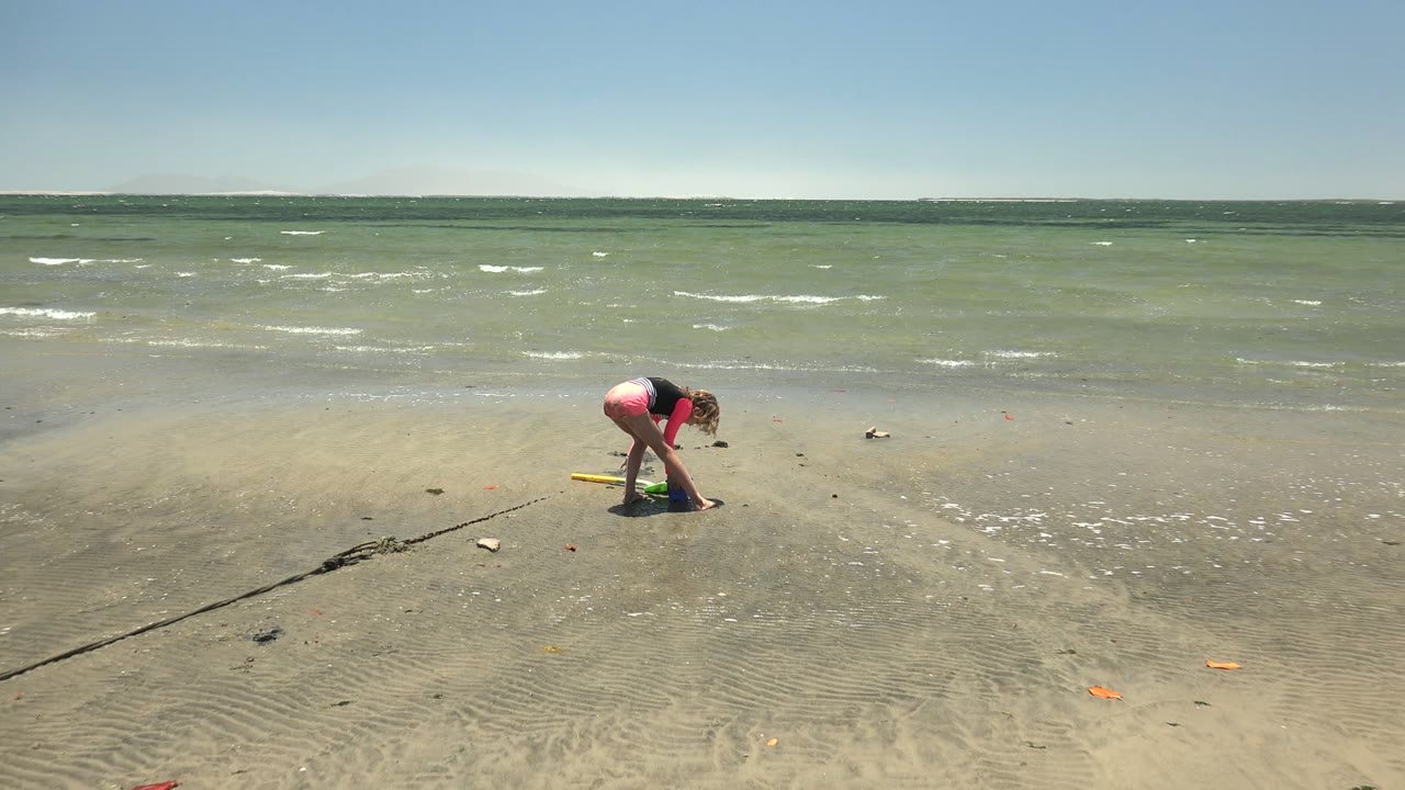 A child in a pink top and blue shorts bends over rippled sand on a San Carlos BCS Mexico beach, with shallow green water and a clear sky behind them.