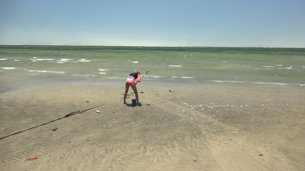 A child on the San Carlos BCS Mexico shoreline stands more upright while holding a green toy, with scattered small beach objects on the sand and wind-textured water behind.