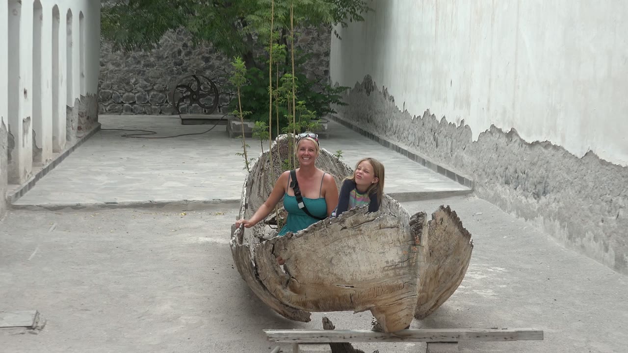 In Mulege BCS Mexico, an adult woman and a child sit side by side inside a large rough wooden bowl in a concrete courtyard with white walls and stonework behind them.