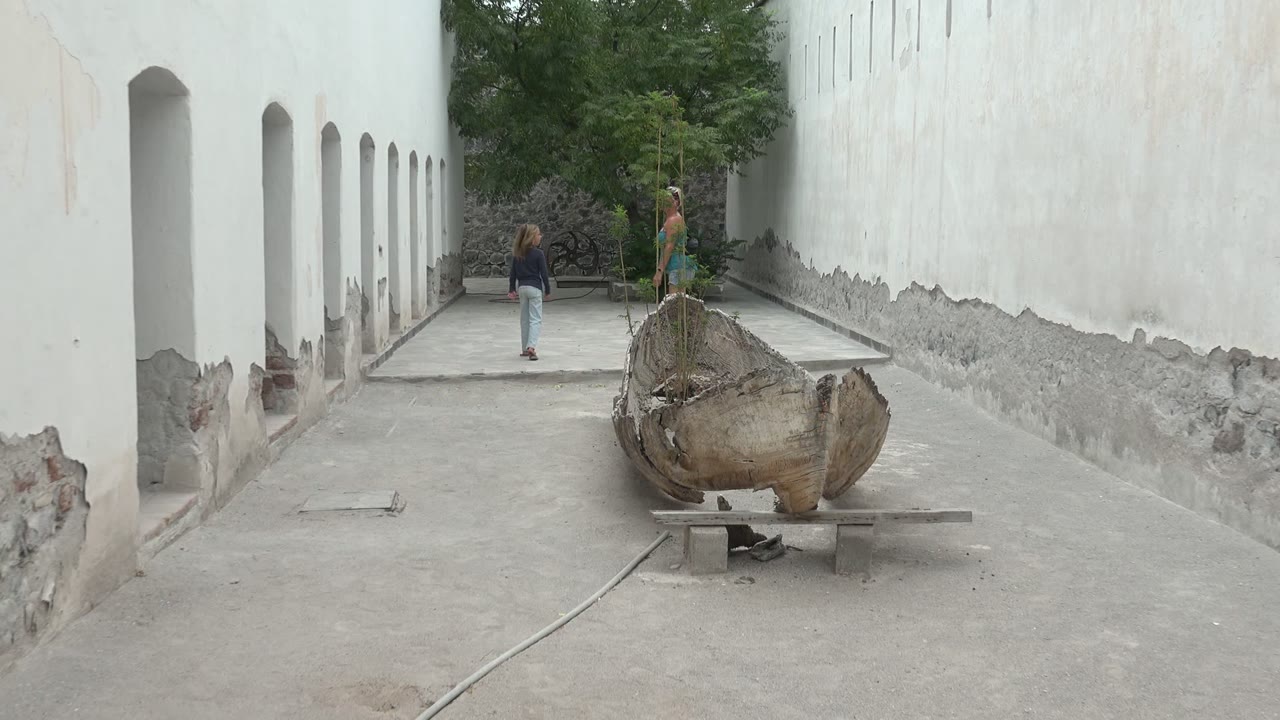 At the start of the Mulege BCS Mexico clip, two people are near the far end of a white-walled courtyard while a weathered wooden object rests on a stand in the foreground.