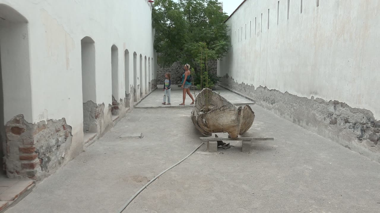 At 0:02, the two people are closer to the camera and pass near the weathered wooden object, with the long white wall and exposed lower stonework running along the right side.