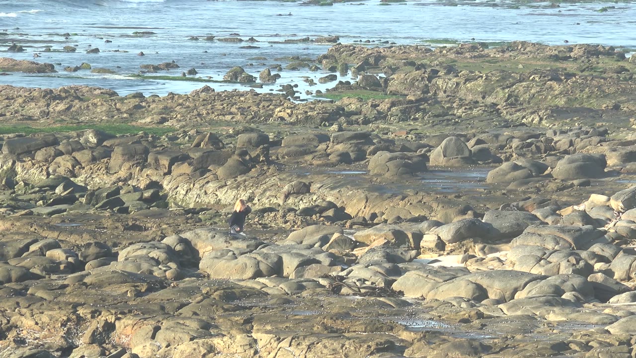 At 0:00 in La Bocana BCS Mexico, a small person stands left of center among rounded coastal rocks with shallow pools and pale ocean water behind them.
