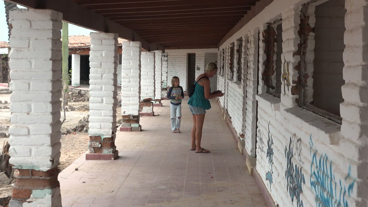 At 0:02, the woman stands near the graffiti-marked wall while the child remains behind her in the shaded Mulege BCS Mexico corridor.