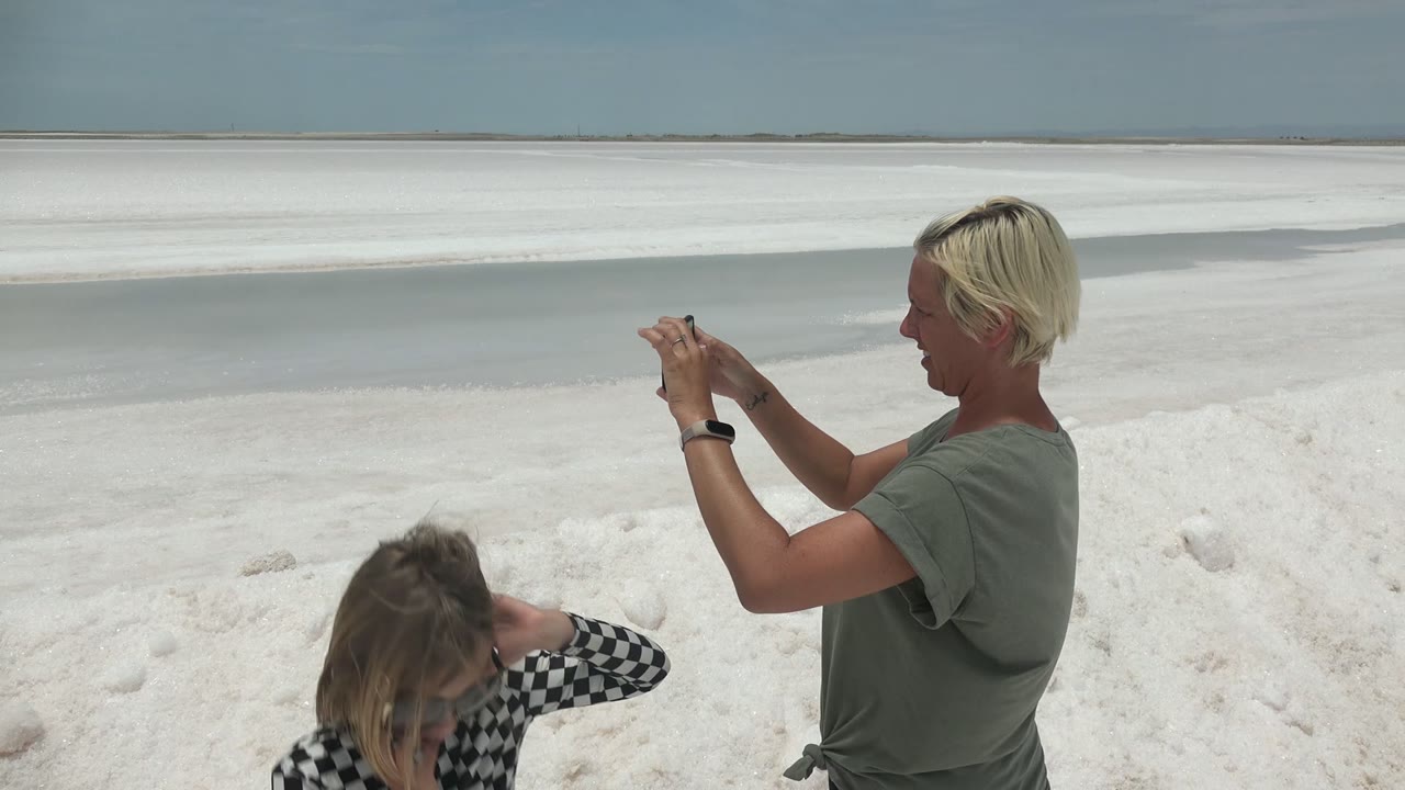 In Guerrero Negro BCS Mexico, a blonde adult holds a smartphone toward a child crouching on a bright white sandy beach beside shallow water.