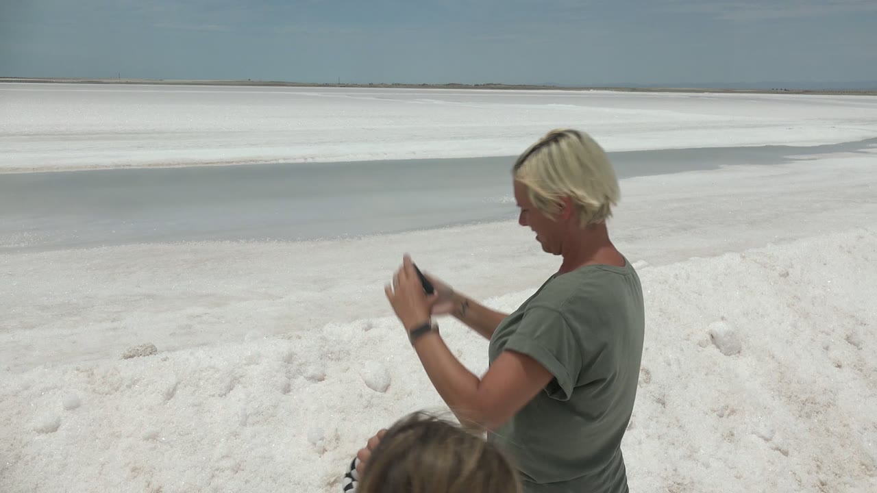 Adult looks at a smartphone while child is partly visible on white sand.