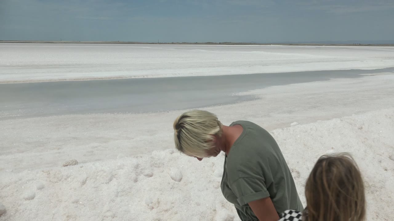 In Guerrero Negro BCS Mexico, the adult bends slightly toward the child near mounded white sand, with shallow water and a flat horizon stretching behind them.
