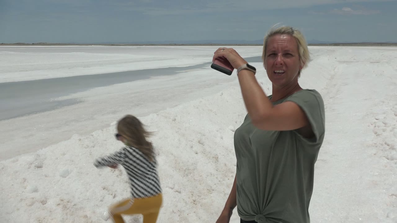 In Guerrero Negro BCS Mexico, a woman in a green shirt holds a phone near her head while another person in yellow pants moves across the white salt flat beside shallow water.
