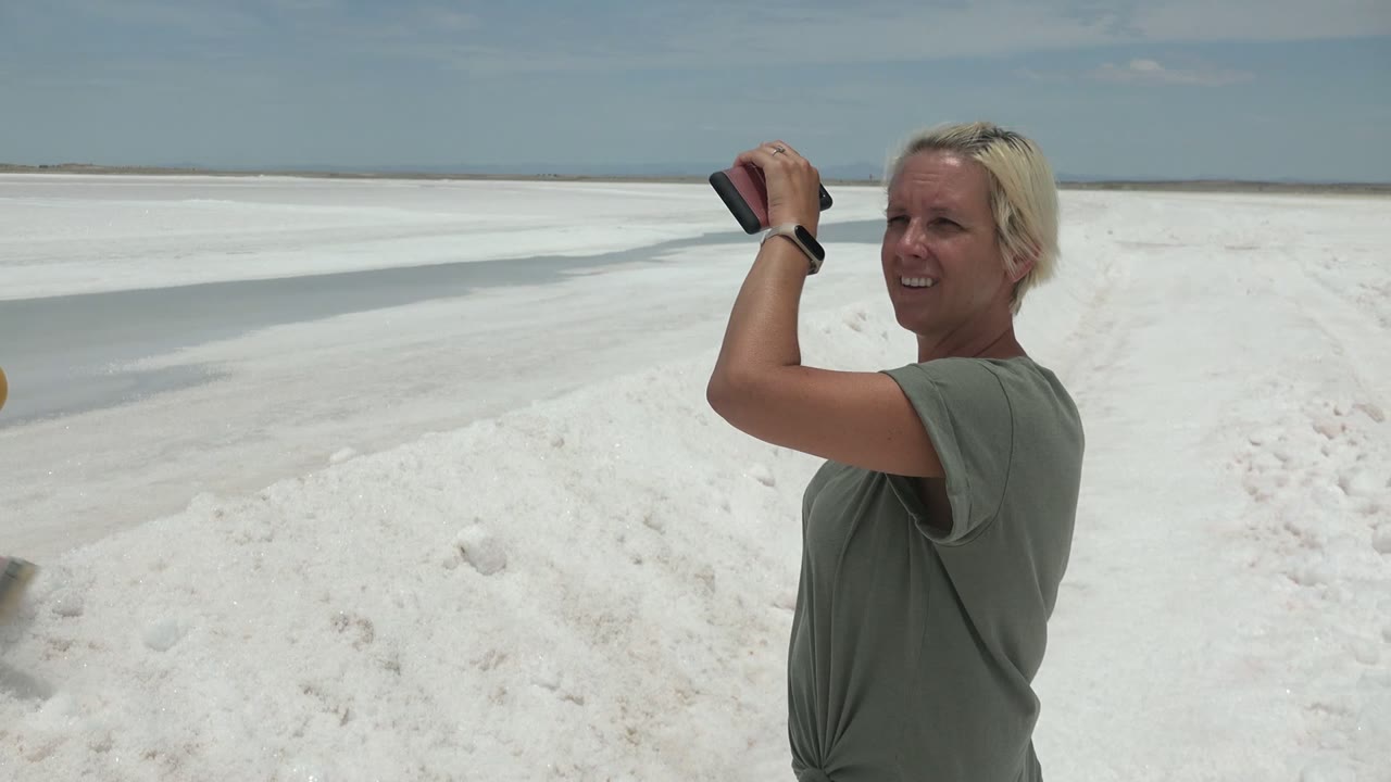 The woman in the green shirt remains at the right side holding a phone, with the second person mostly out of frame and the white salt flat stretching to the horizon in Guerrero Negro BCS Mexico.