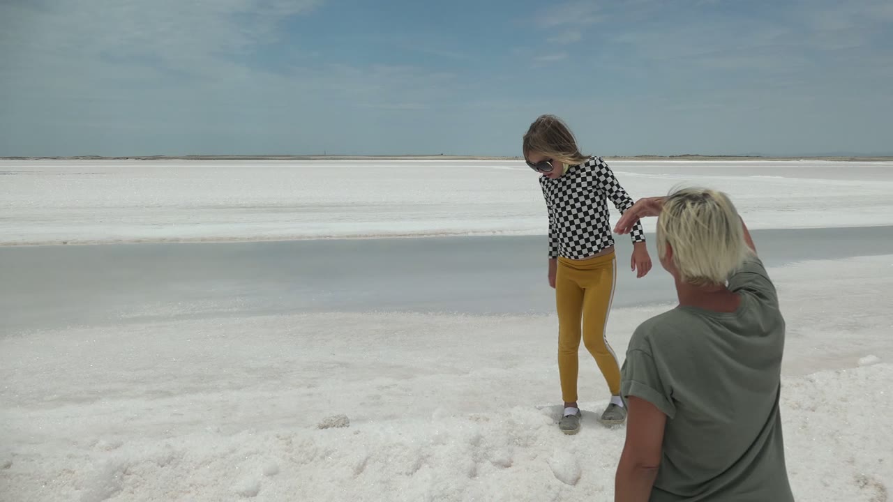 In Guerrero Negro BCS Mexico, an adult in a gray shirt faces away from camera while a child in a checkered shirt and yellow pants stands on the white salt flat under blue sky.