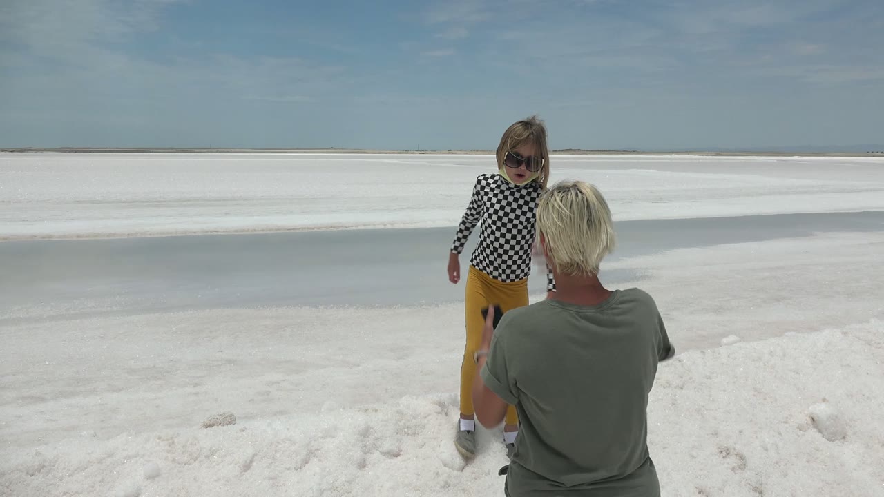The adult's back fills more of the foreground while the child stands to the left on the white salt flat in Guerrero Negro BCS Mexico, with pale waterlike bands across the background.