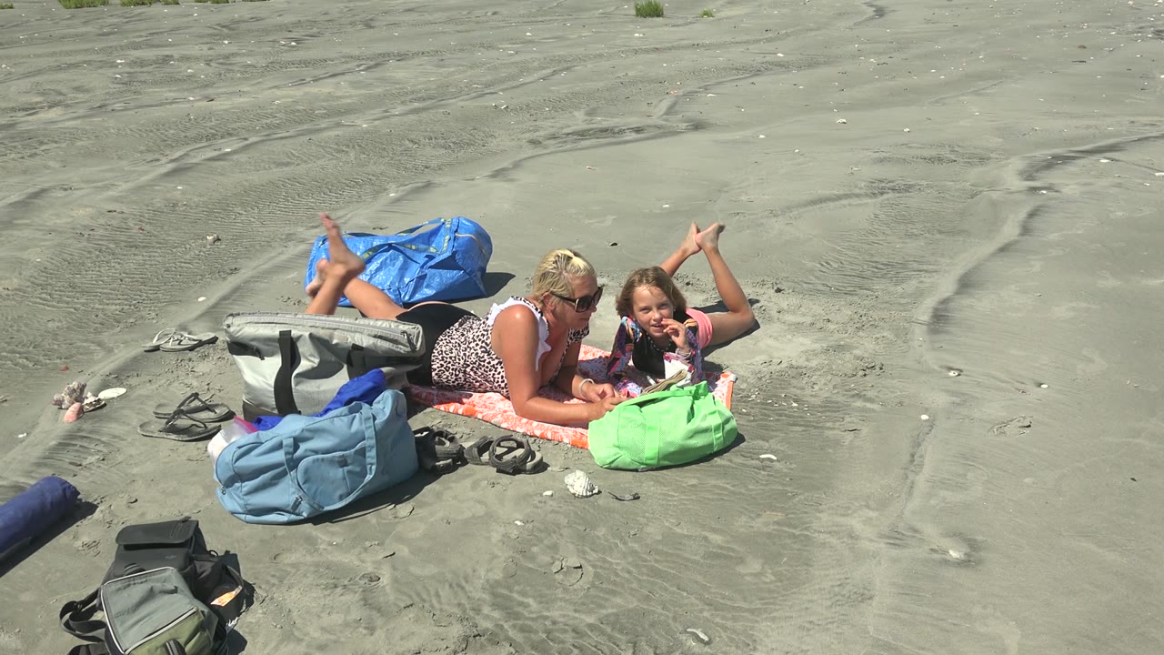 At La Bocana BCS Mexico, an adult and child lie on an orange towel beside blue, green, and gray bags while reading on open gray sand.
