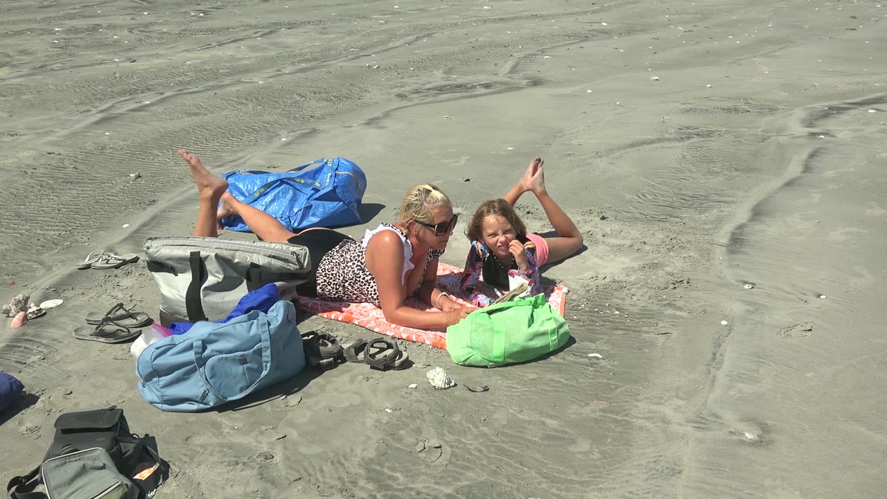 Adult and child reading together beside colorful bags on the beach.