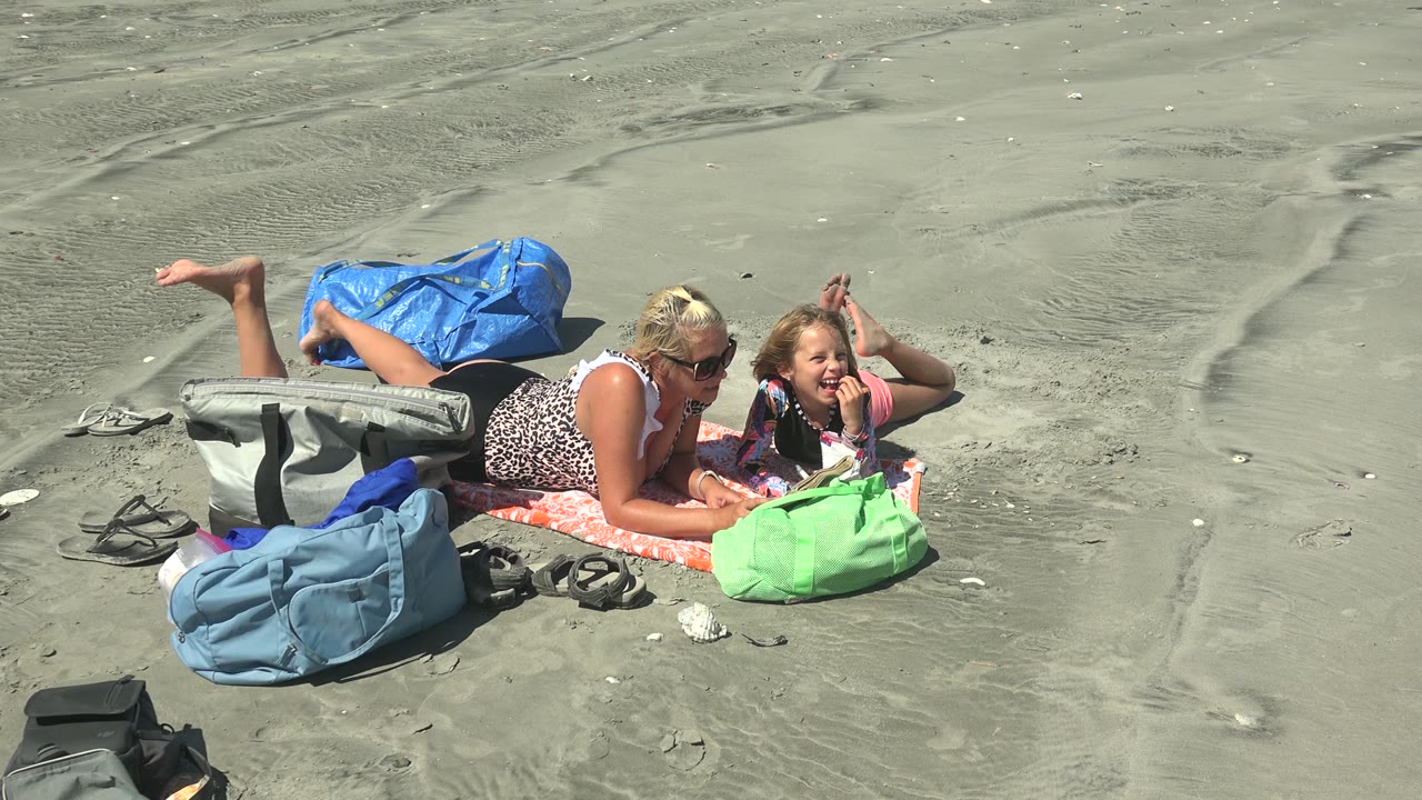 At La Bocana BCS Mexico, the child faces upward with a visible smile while the adult remains beside the open book and beach bags on gray sand.
