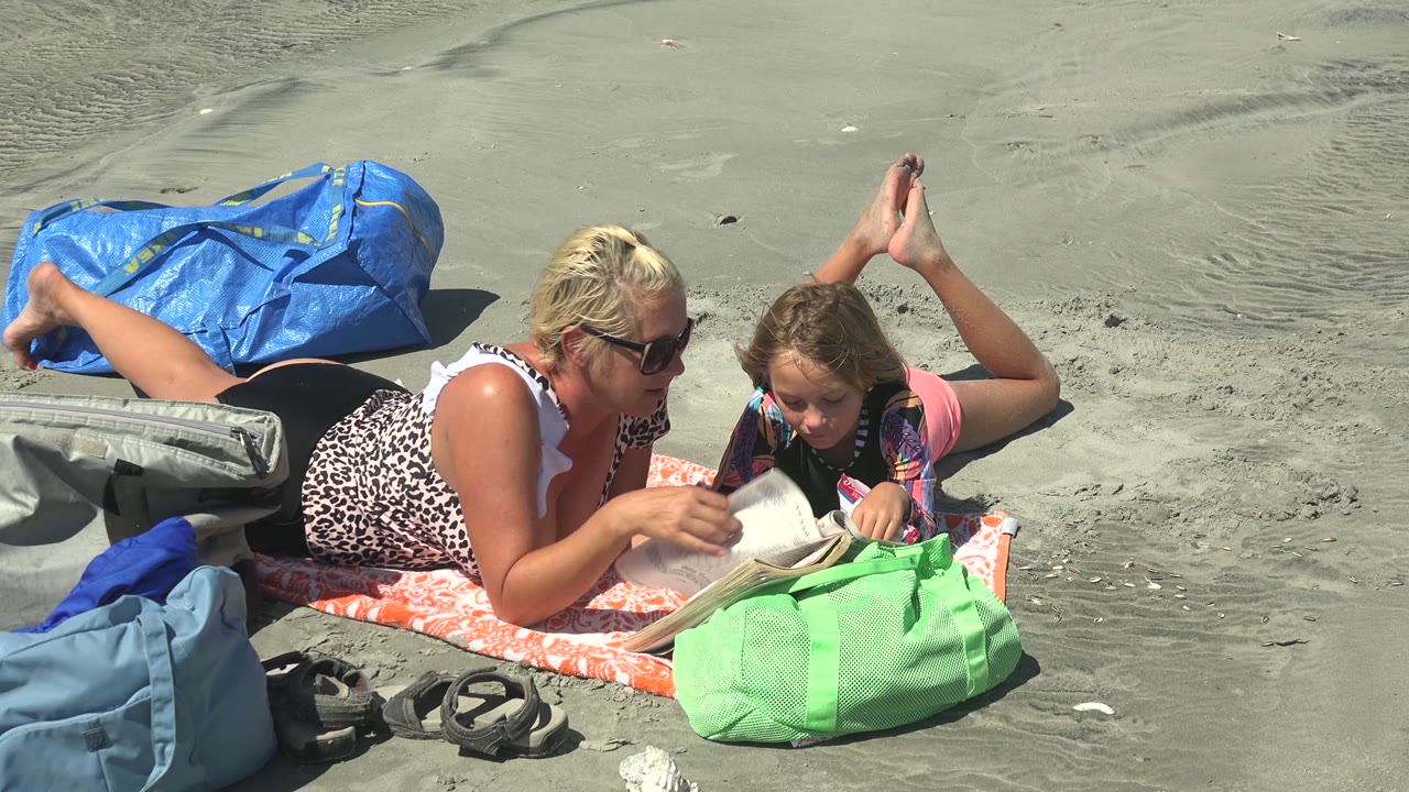 At La Bocana BCS Mexico, a woman in sunglasses and a child lie on an orange towel while reading a book, with a green bag in the foreground and a blue bag behind them.