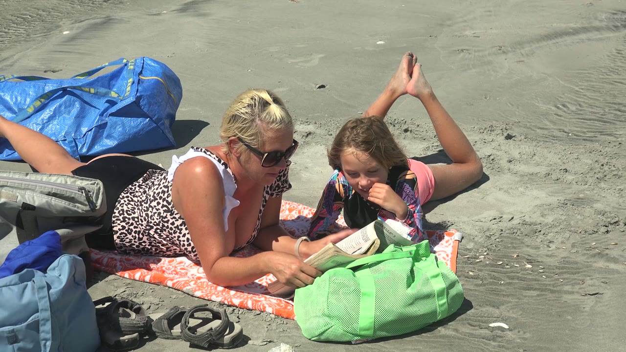 Woman points near an open book as a child watches on a beach towel.