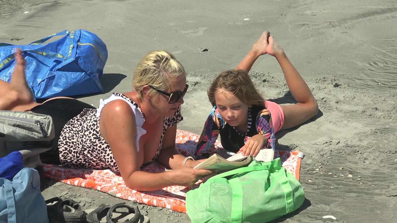 At La Bocana BCS Mexico, the child bends over the book while the woman holds or steadies the pages, with a blue bag behind them and beach gear clustered nearby.