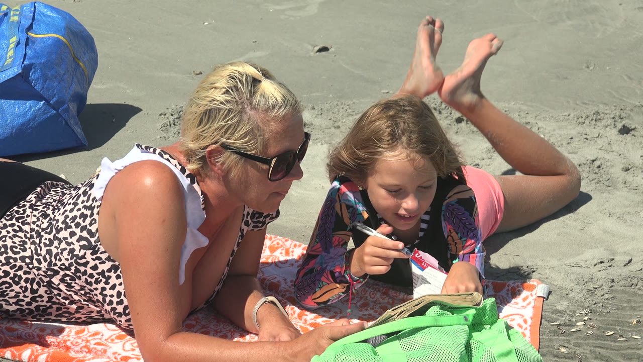 At 0:00 in La Bocana BCS Mexico, a woman in sunglasses lies beside a child holding a pen over a book on an orange towel, with green and blue bags on the sand.