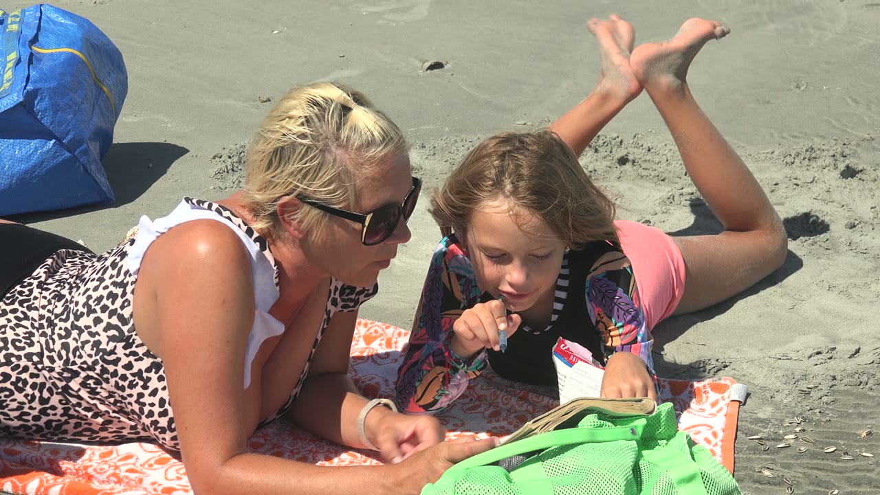 At 0:02 in La Bocana BCS Mexico, the woman extends a hand near the towel while the child keeps the pen near their mouth and the book remains open on the sand.