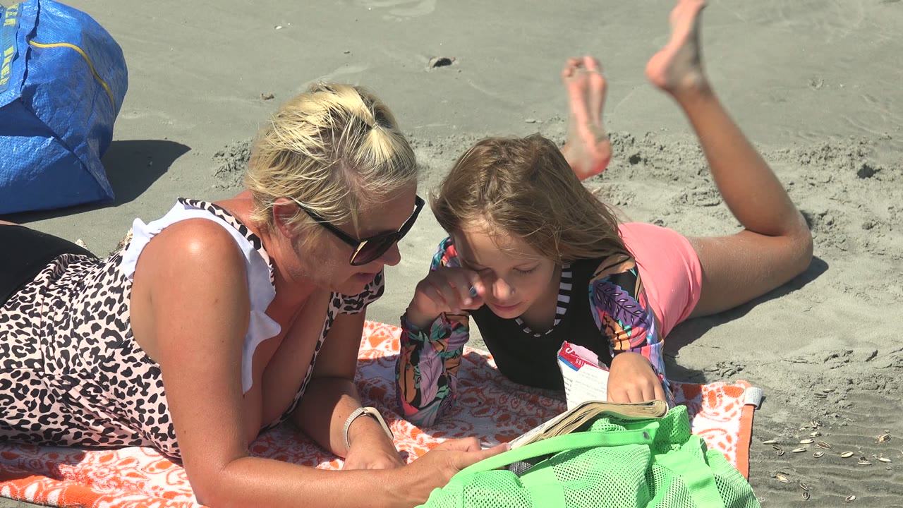 At La Bocana BCS Mexico, a woman in sunglasses and a child lie on an orange towel on gray sand with a book open in front of them and a green bag in the foreground.