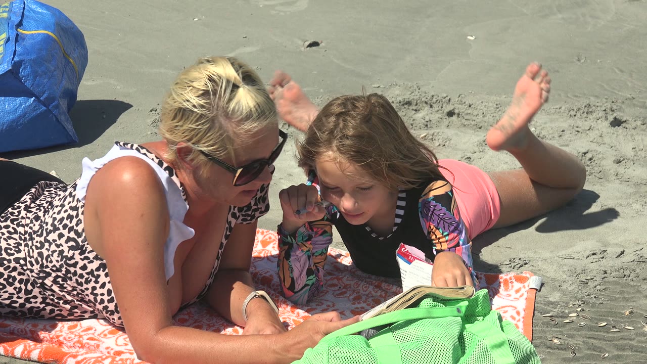 Woman watches a child look down at an open book on a beach towel.