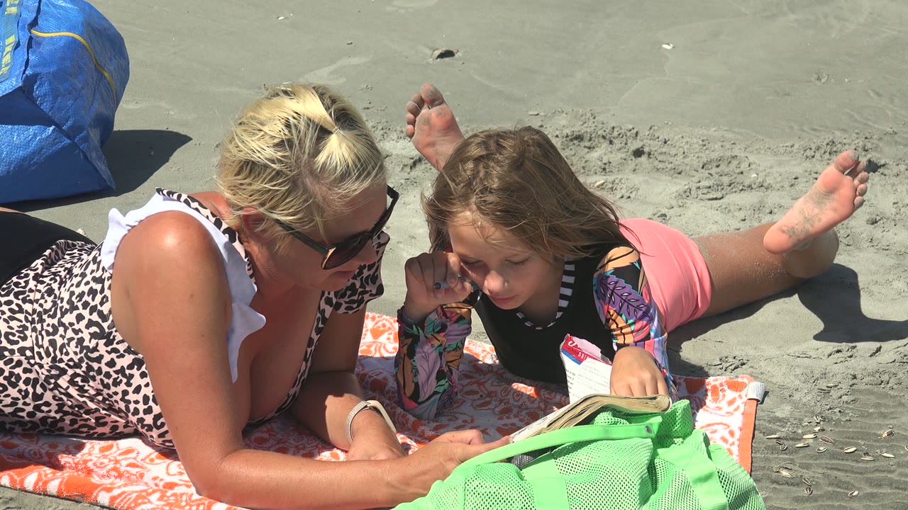 At La Bocana BCS Mexico, the child raises a hand near her face while lying beside the woman on the towel, with the open book and green bag still in the foreground.