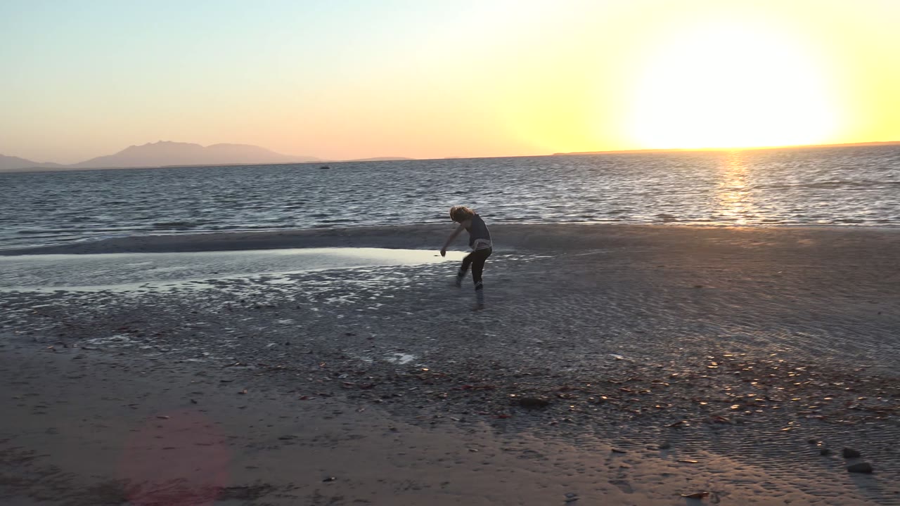 In San Carlos BCS Mexico, a child bends near the wet sand while the low sun reflects on the ocean and distant mountains sit along the horizon.
