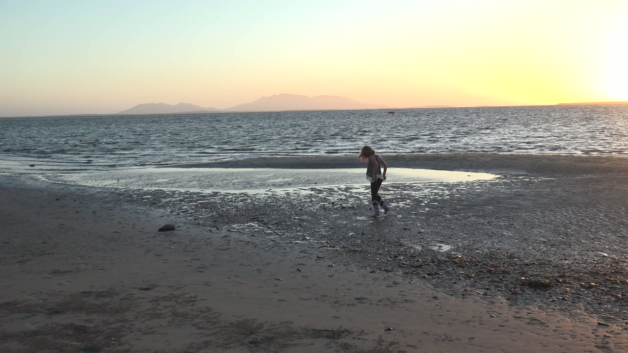 In San Carlos BCS Mexico, the child moves past a shallow reflective pool on the beach while the low sun brightens the right side of the ocean horizon.