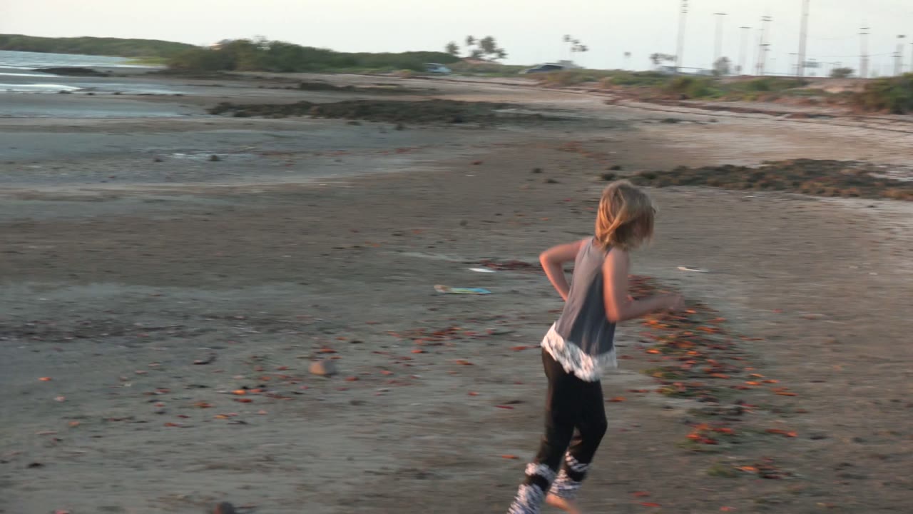 The child runs farther along the beach in San Carlos BCS Mexico, with darker wet sand giving way to drier shore and low structures far to the right.