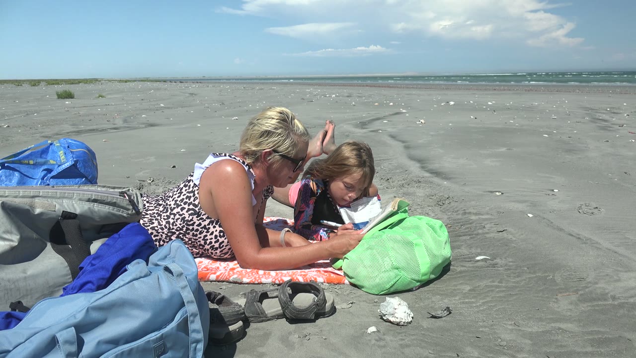 A woman and child read together on an orange towel at La Bocana BCS Mexico, with a green bag, blue beach gear, gray sand, and distant surf under bright sky.