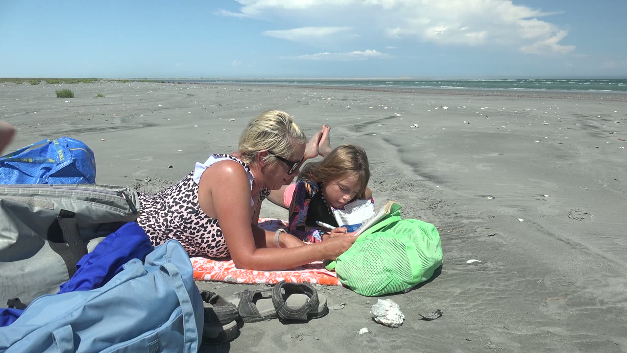 Woman holds a book while a child reads beside beach bags.